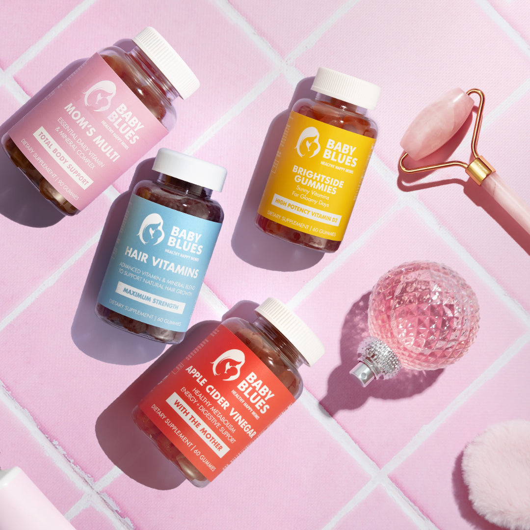 Baby Blues Hair Vitamins, Mom’s Multi, Brightside Gummies, and ACV Gummies arranged on pink tiles with beauty tools