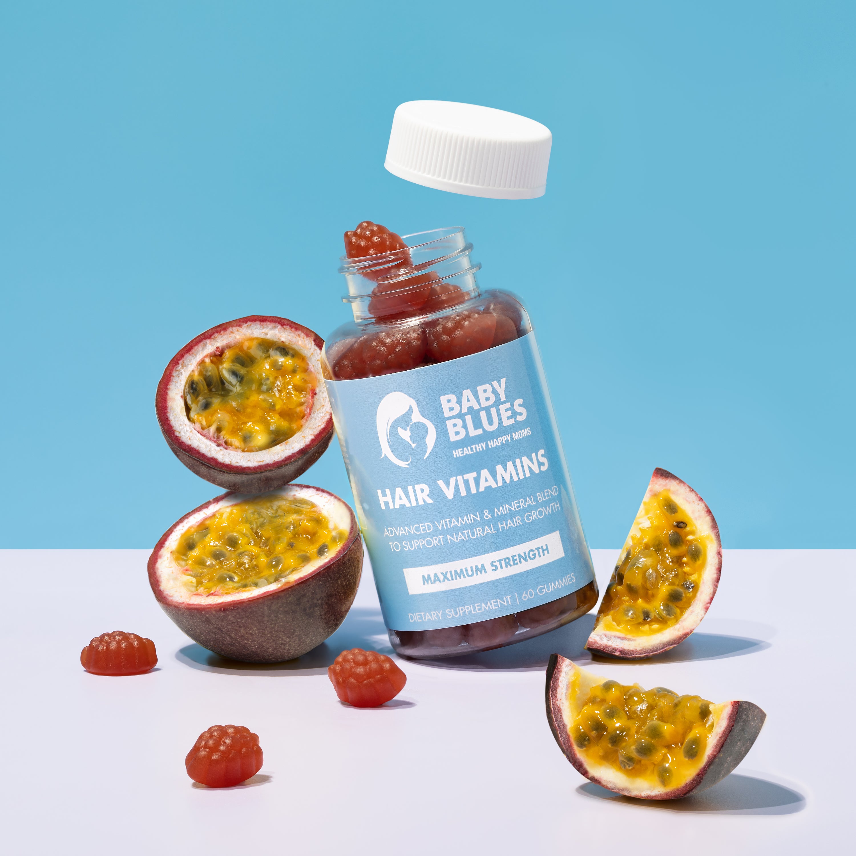 Baby Blues Hair Vitamins bottle surrounded by sliced passion fruit and gummies on a bright blue backdrop.