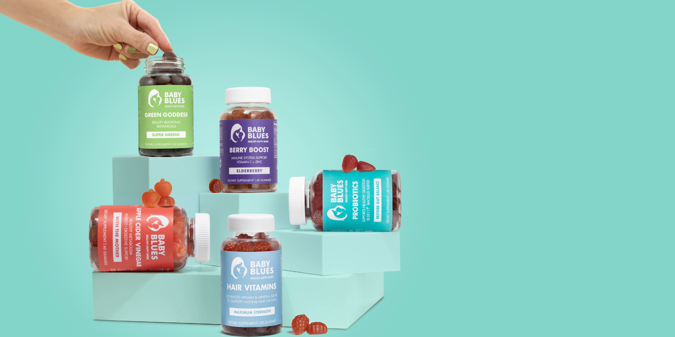 Hand lifting Baby Blues Green Goddess bottle beside full supplement lineup displayed on pastel blocks.